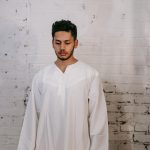 A young man in a thobe stands against a textured brick wall, exuding serenity and tradition.