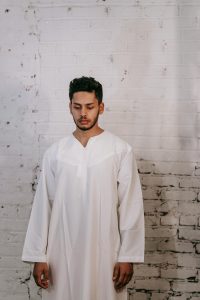 A young man in a thobe stands against a textured brick wall, exuding serenity and tradition.